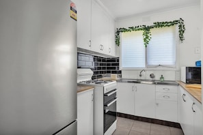 A kitchen with white cabinets, a stove, an oven, a microwave, and a window with blinds.