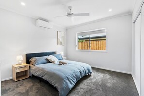 Ein Schlafzimmer mit einem Bett, einem Nachttisch, einem Deckenventilator, einer Klimaanlage, einem Fenster mit Blick auf einen Zaun und einem gerahmten Bild an der Wand.