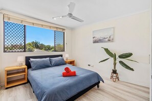 A bedroom with a bed, a ceiling fan, a window with a view, a bedside table, a nightstand, and a potted plant. - Sunny, Stylish Island Stay Minutes from Surfers (Surfers Paradise)