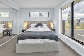 A hotel room with a large bed, two bedside tables with lamps, a view of the outside through a large window, and a modern headboard. - Bright and Cozy 4-Bed Home in Rockbank (Rockbank)
