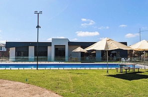 A swimming pool with a modern building in the background, outdoor seating with umbrellas, and a clear sky. - Bright and Cozy 4-Bed Home in Rockbank (Rockbank)