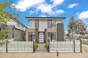 A modern two-story house with a white picket fence, a driveway, and a clear blue sky. - Bright and Cozy 4-Bed Home in Rockbank (Rockbank)