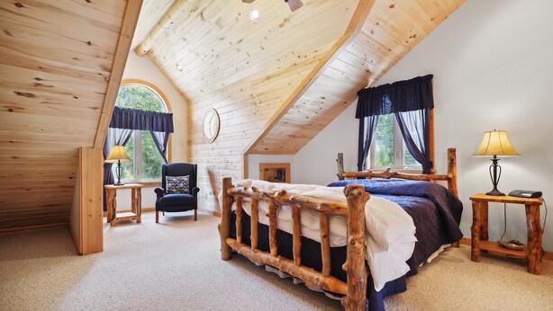A bedroom with a wooden ceiling, a bed with a rustic headboard, a chair, two lamps, and a window with curtains.