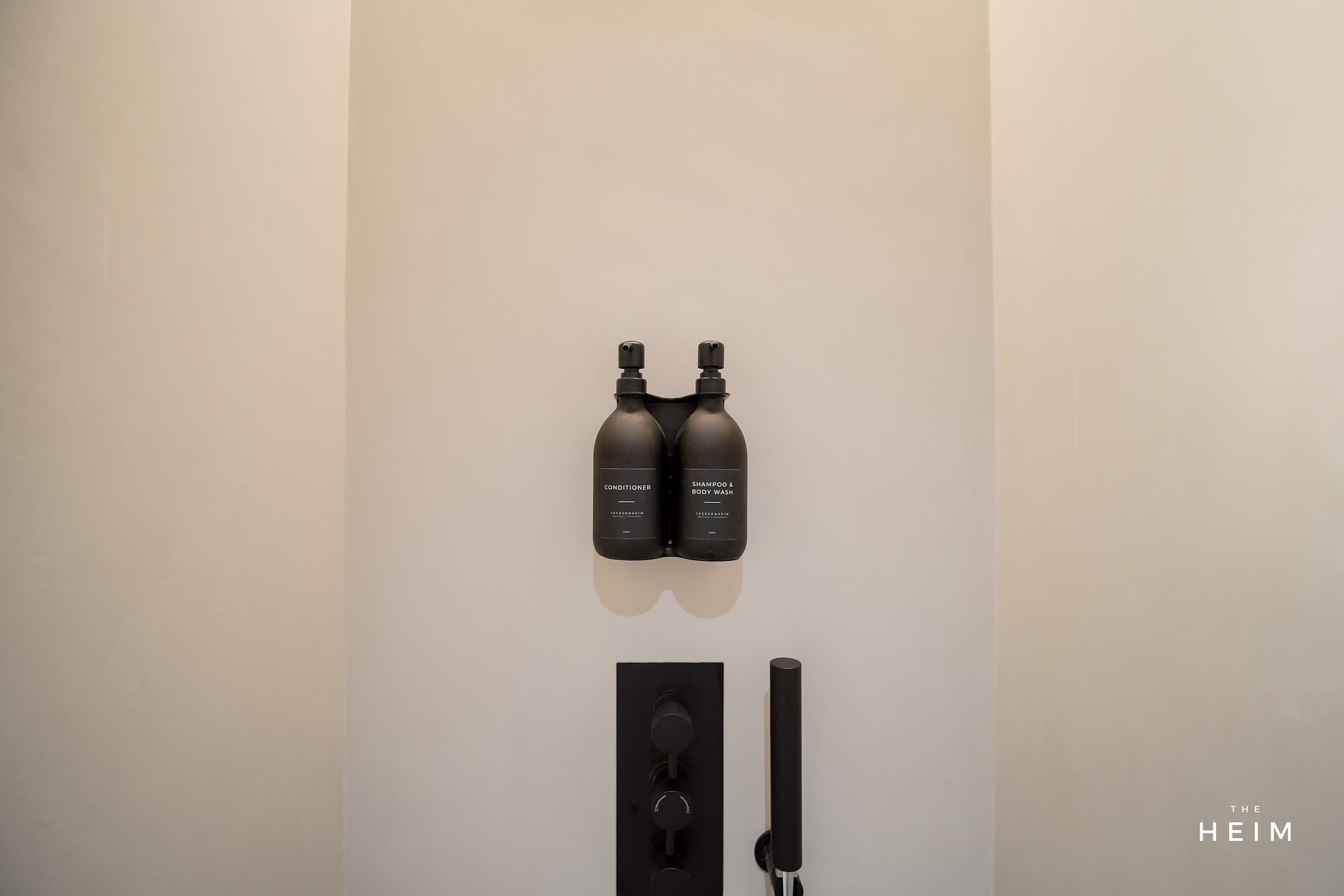 A bathroom with two bottles labeled "Conditioner" and "Shampoo & Body Wash" on the wall, a faucet, and a control panel.