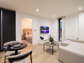 Ein modernes Wohnzimmer mit einer Couch, einem Esstisch mit Stühlen, einem an der Wand montierten Fernseher und einem Bett im angrenzenden Zimmer.