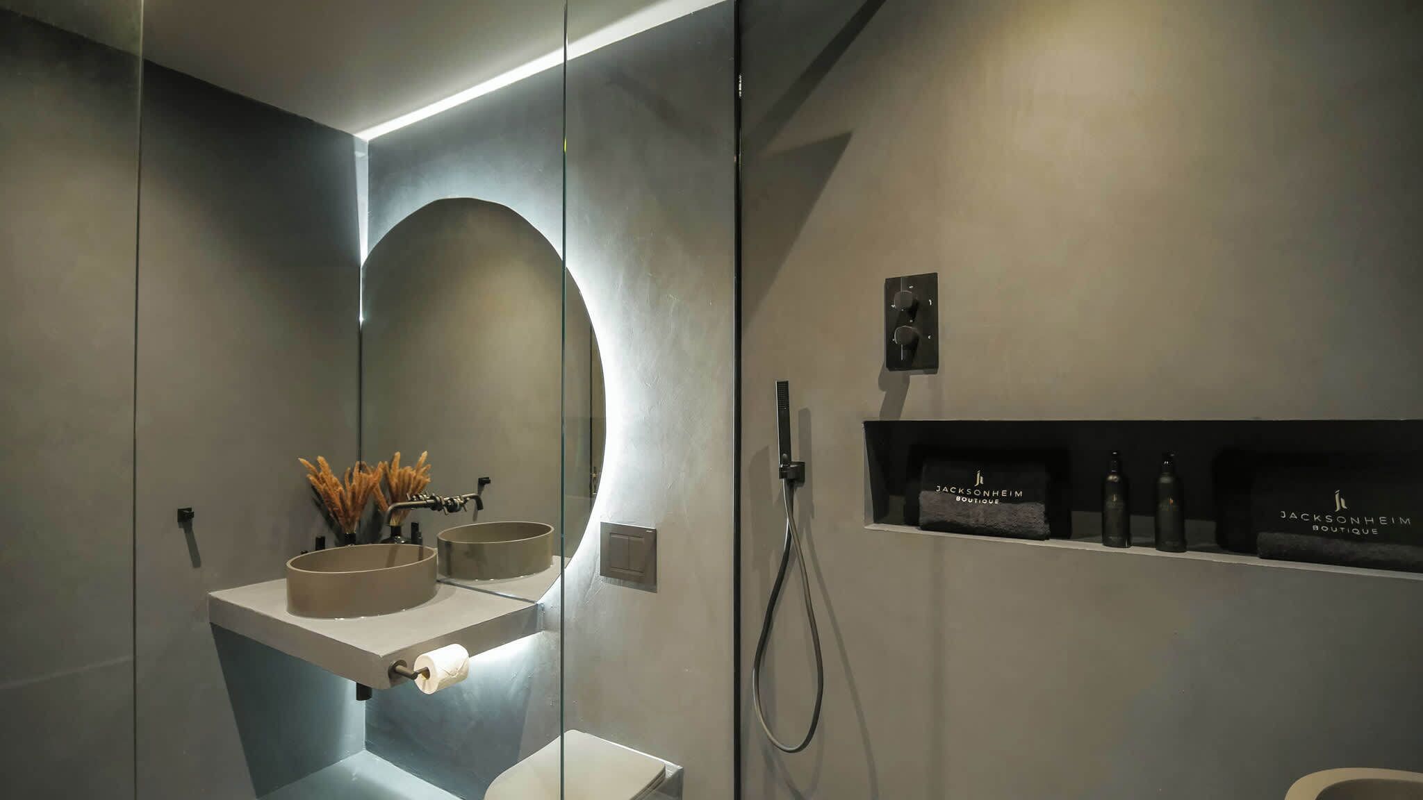A modern bathroom with a large mirror, a sink with two basins, a shower, and a shelf with toiletries.