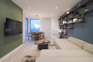 A modern living room with a flat-screen TV, a sofa, a dining area with a table and chairs, and shelves with decorative items. - Midori | The Heim (Manchester)