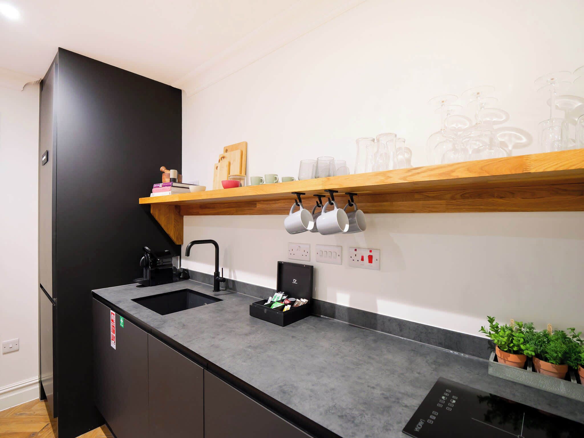 A kitchen with a black refrigerator, a sink, a wooden shelf with mugs, and a stove.