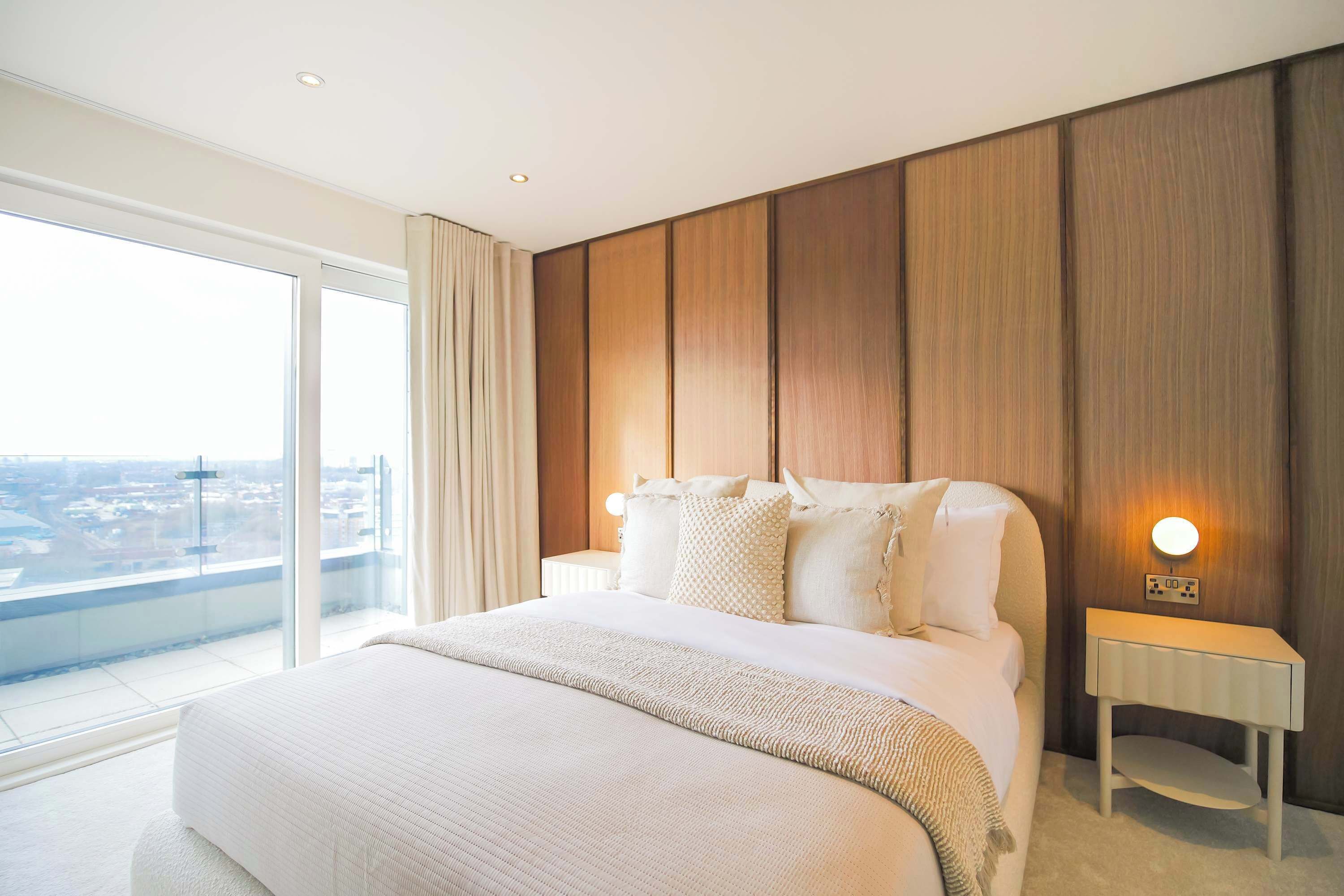 Een moderne hotelkamer met een groot bed, een nachtkastje, een wandlamp en een groot raam met uitzicht op de stad.