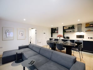 Ein modernes Wohnzimmer mit einem grauen Ecksofa, einem kleinen runden Tisch und einem Essbereich mit einem weißen Tisch, gedeckt für vier Personen.