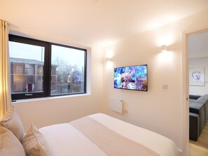 Ein Schlafzimmer mit Bett, einem an der Wand montierten Fernseher, einem Fenster mit Blick auf Gebäude und einer Tür zu einem anderen Raum.