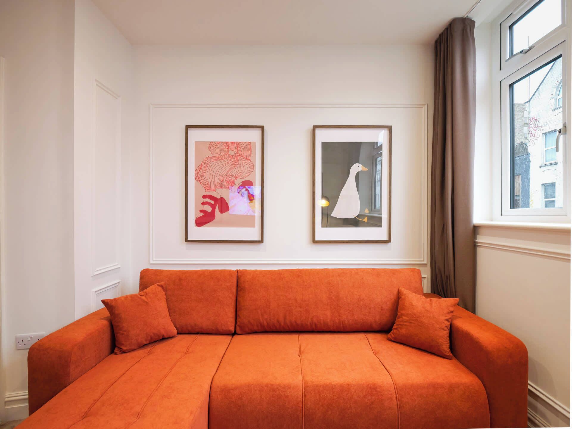 Ein Zimmer mit einer Couch, zwei gerahmten Kunstwerken, einem Fenster mit Vorhängen und Blick auf Gebäude.