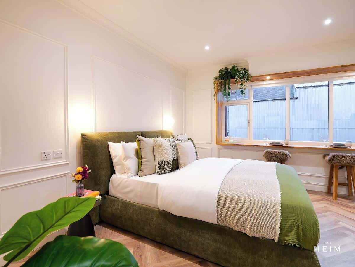 A bedroom with a large bed, a window with blinds, a small stool, and a plant.