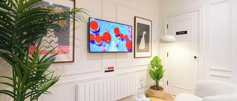A modern living room with a flat-screen TV, framed artwork, a white door, a plant, a round coffee table with books and a bowl, and a white sofa.