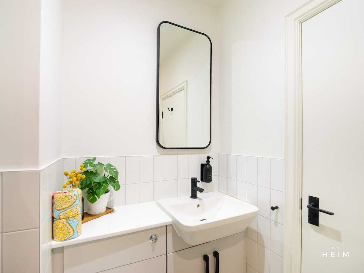 A bathroom with a white sink, black faucet, and a mirror above it. There is a potted plant on the counter and a roll of toilet paper.