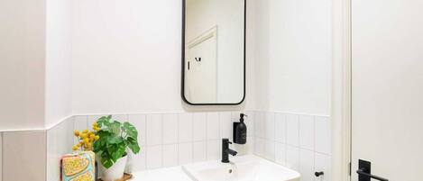A bathroom with a white sink, black faucet, and a mirror above it. There is a potted plant on the counter and a roll of toilet paper.