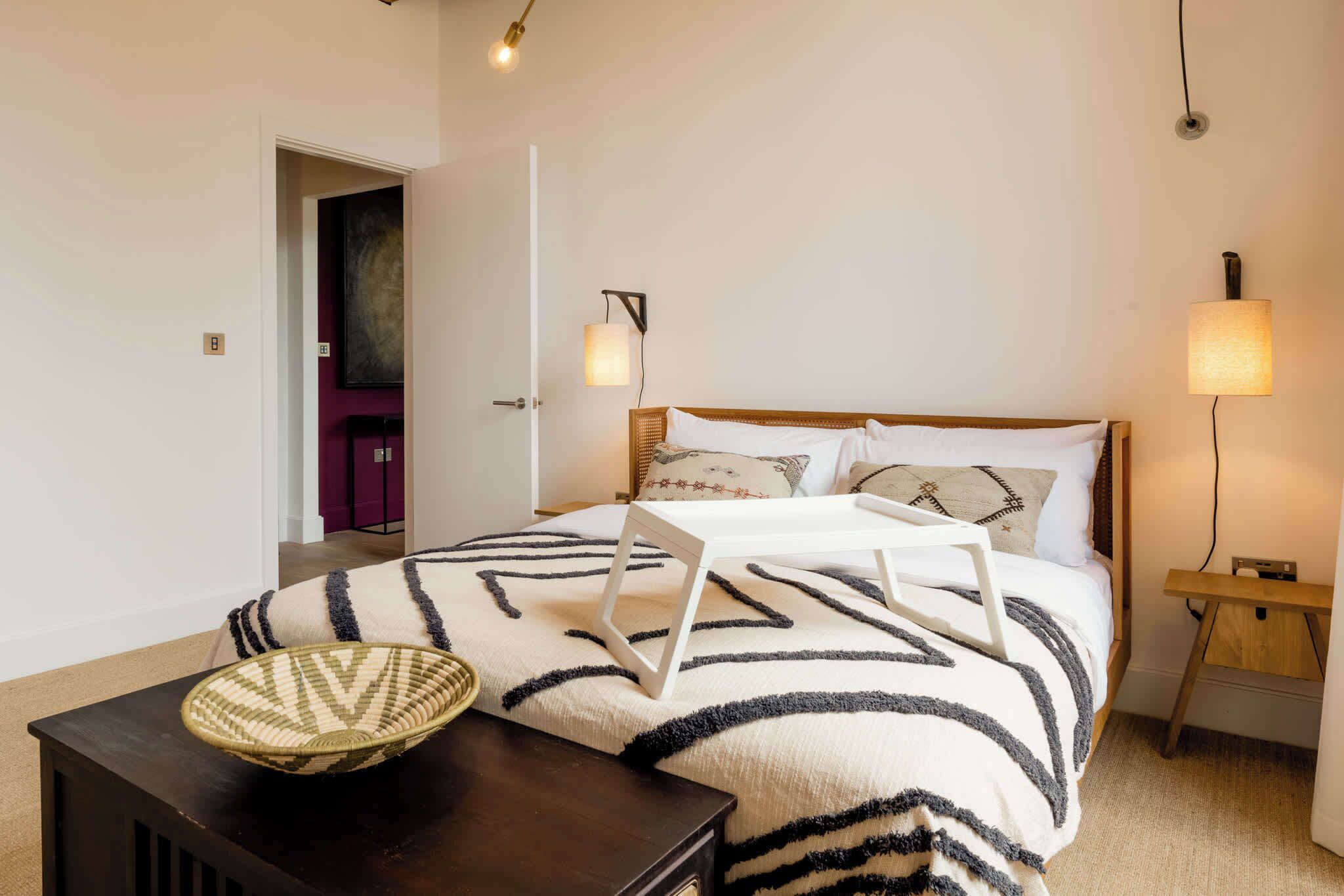 A bedroom with a bed, bedside lamps, a nightstand, a woven basket, and a door leading to another room.