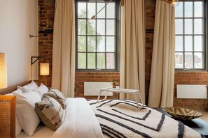 A bedroom with a brick wall, large windows with curtains, a bed with pillows, a bedside table, and a floor lamp. - Africana | The Heim (Manchester)