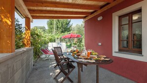 Un'area pranzo all'aperto con un tavolo apparecchiato per un pasto, circondato da sedie e un ombrellone rosso.