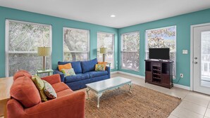 Una sala de estar con paredes verde azulado, un sofá azul, un sillón rojo, una mesa de centro blanca y un mueble para televisión.