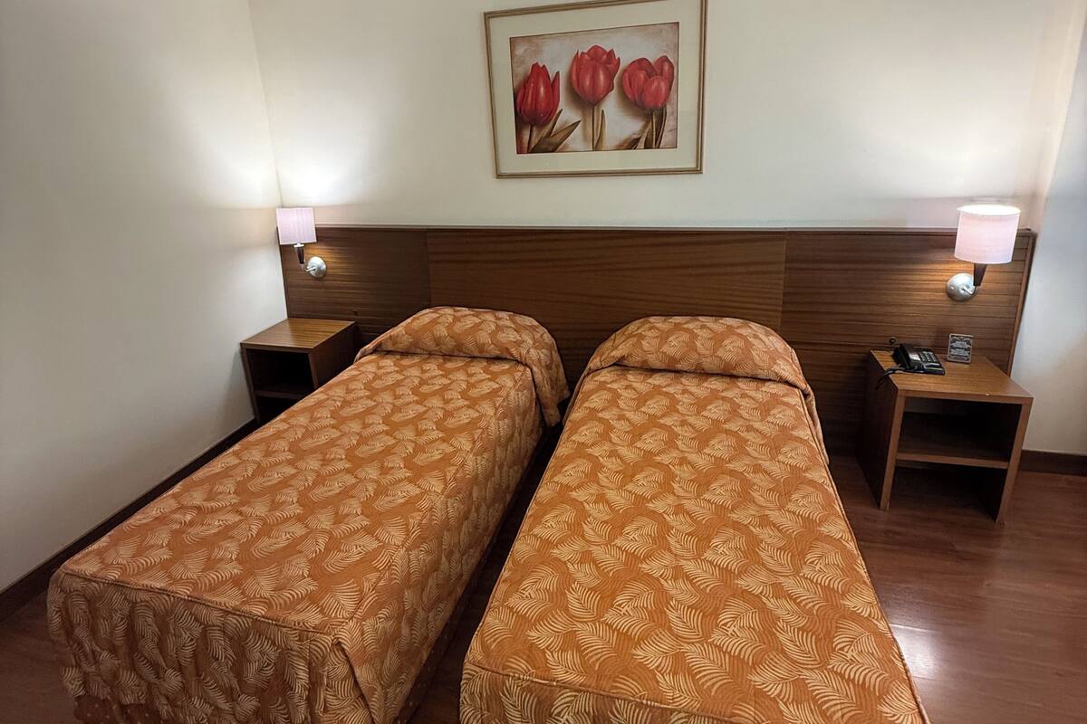 Habitación de hotel con dos camas individuales, cabecero de madera, mesitas de noche con lámparas y un cuadro en la pared.