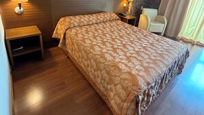Hotellihuone, jossa on kuviollinen päiväpeite, kaksi yöpöytävalaisinta, puinen yöpöytä, tuoli ja kuvioitu seinä.