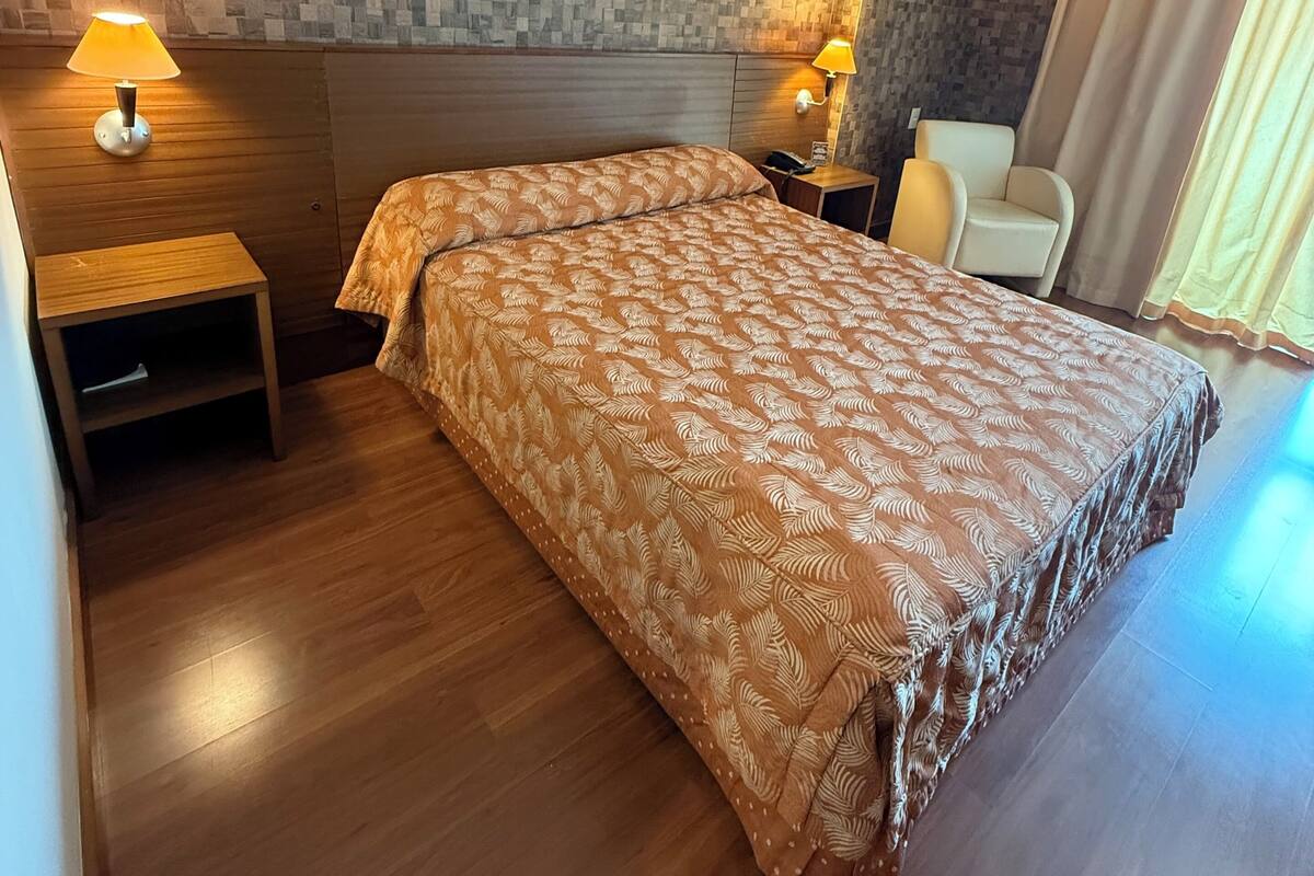 Habitación de hotel con una colcha estampada, dos lámparas de noche, una mesita de madera, una silla y una pared con textura.