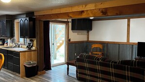 A cozy living room with a wooden floor, a plaid sofa, and a kitchen area with dark cabinets and a wooden ceiling. - Tobacco Riverwalk Condo - Eureka, MT (Eureka)