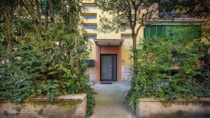 Une entrée d’immeuble avec une porte, entourée de verdure et bordée d’un chemin en béton.
