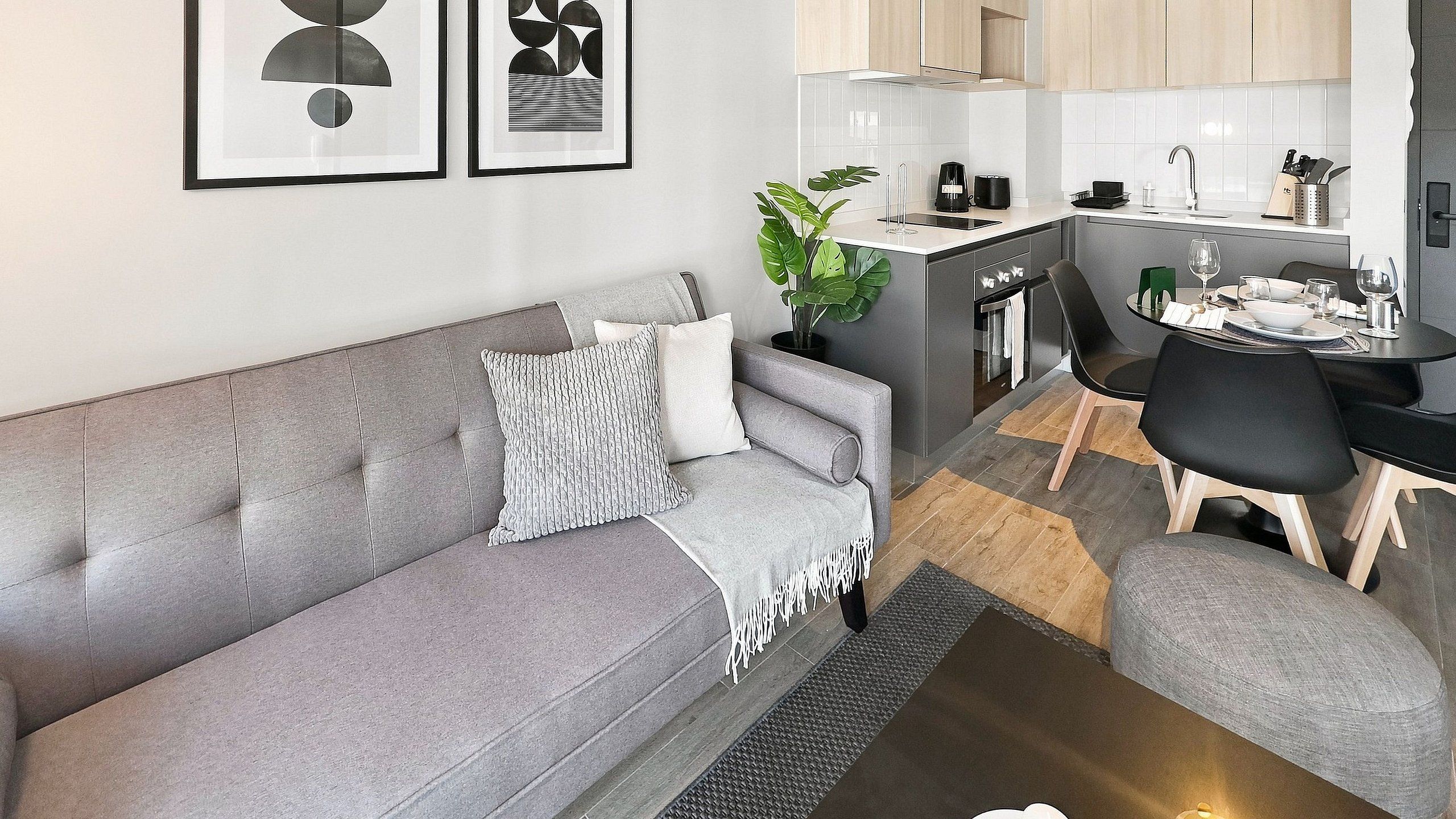 En moderne stue med en grå sofa, et spisebord med stole, et køkken og en potteplante.