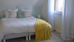 Ein Schlafzimmer mit einem Bett, weißer Bettwäsche, einer gelben Tagesdecke und einem Fenster mit durchsichtigen Vorhängen.