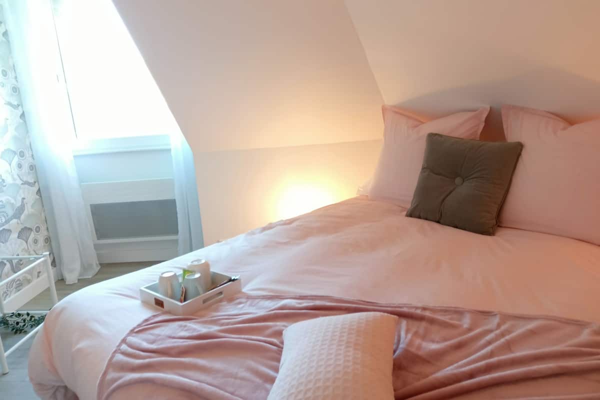 Une chambre à coucher avec un lit, des oreillers, une table de chevet sur laquelle se trouve une petite table avec des articles de toilette, et une fenêtre avec des rideaux.