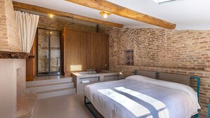 Une chambre avec un grand lit, un plafond en bois, un mur en pierre et une fenêtre avec des rideaux.
