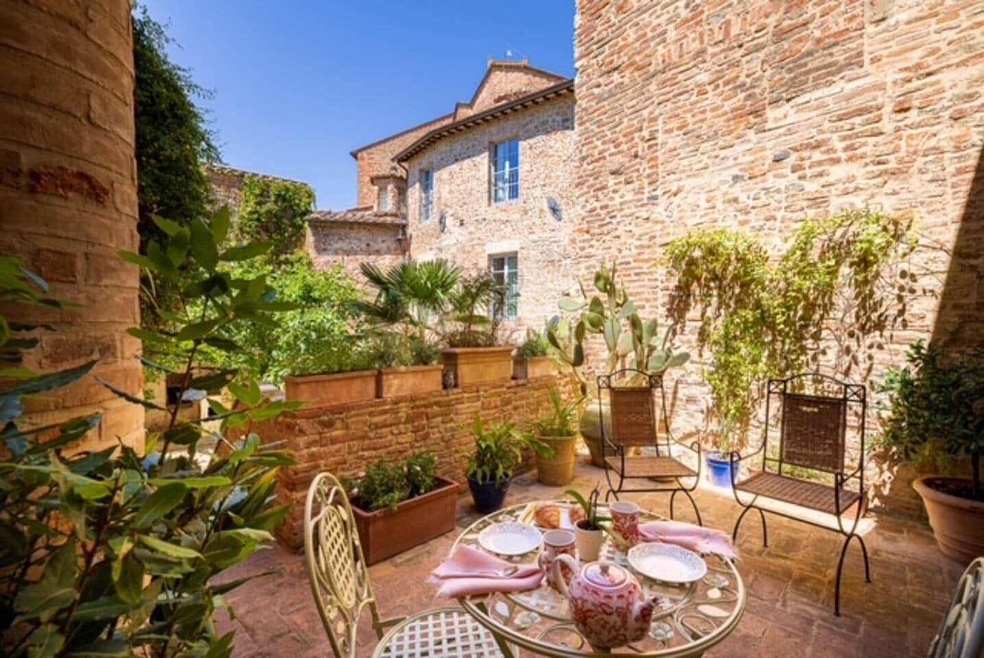 A courtyard with a table set for dining, surrounded by potted plants and stone walls.