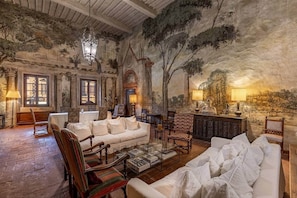 Un vaste salon doté d’un haut plafond, de colonnes richement décorées et d’une fresque représentant un paysage naturel. On y trouve plusieurs coins salon avec des canapés blancs et des fauteuils à motifs.