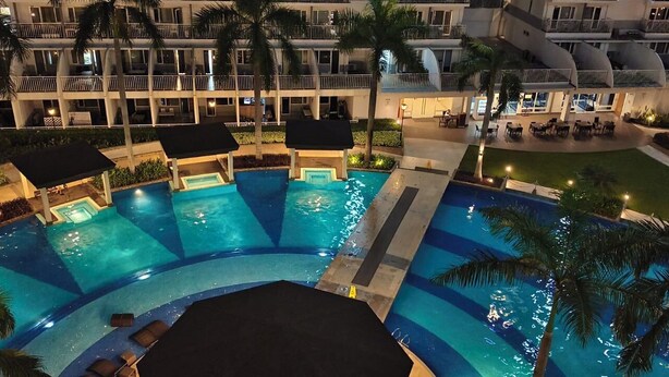 A multi-level hotel with a large, curved swimming pool surrounded by palm trees and lounge areas. - MOA Staycation Amenity view Escape @ Shell residence (Pasay City)