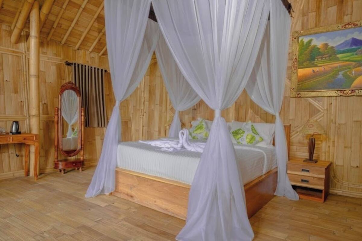 Een houten kamer met een bed onder een baldakijn, een houten nachtkastje, een spiegel en een schilderij aan de muur.
