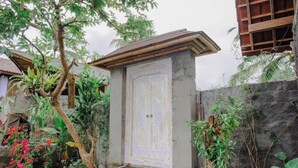 Une maison traditionnelle en bois, dotée d’une porte en pierre, entourée d’une végétation luxuriante et d’un mur de pierres.