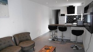 Eine moderne Küche mit weißen Schränken, schwarzen Arbeitsplatten und einer schwarzen Spritzschutzwand. Es gibt drei Barhocker und einen Holztisch mit einer Fernbedienung darauf.