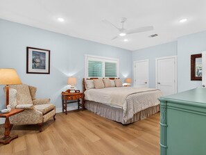 Une chambre à coucher comprenant un lit, des tables de chevet, une chaise, un ventilateur de plafond et une fenêtre avec des stores.