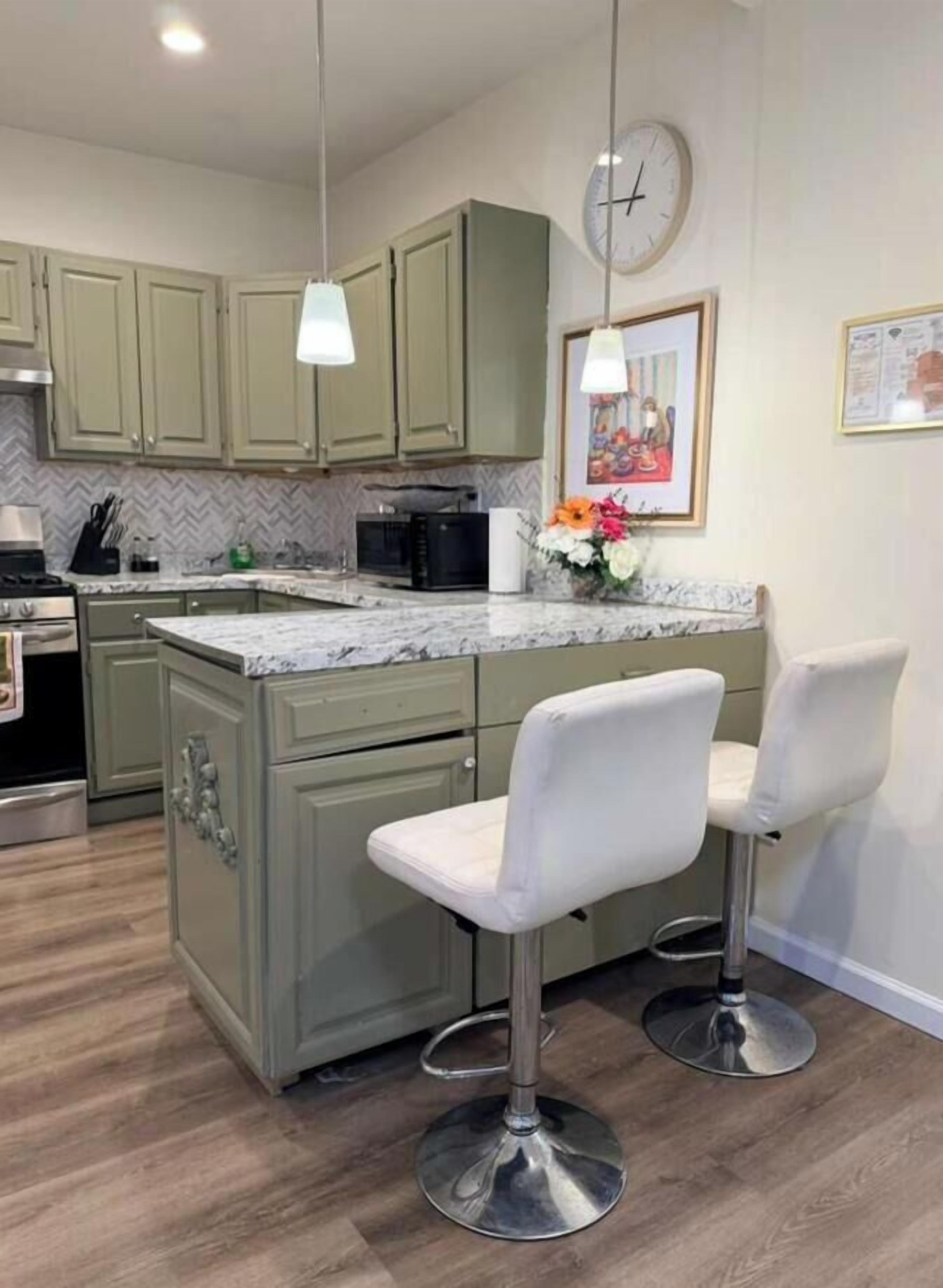 A kitchen with green cabinets, a marble countertop, a clock, and two white bar stools.