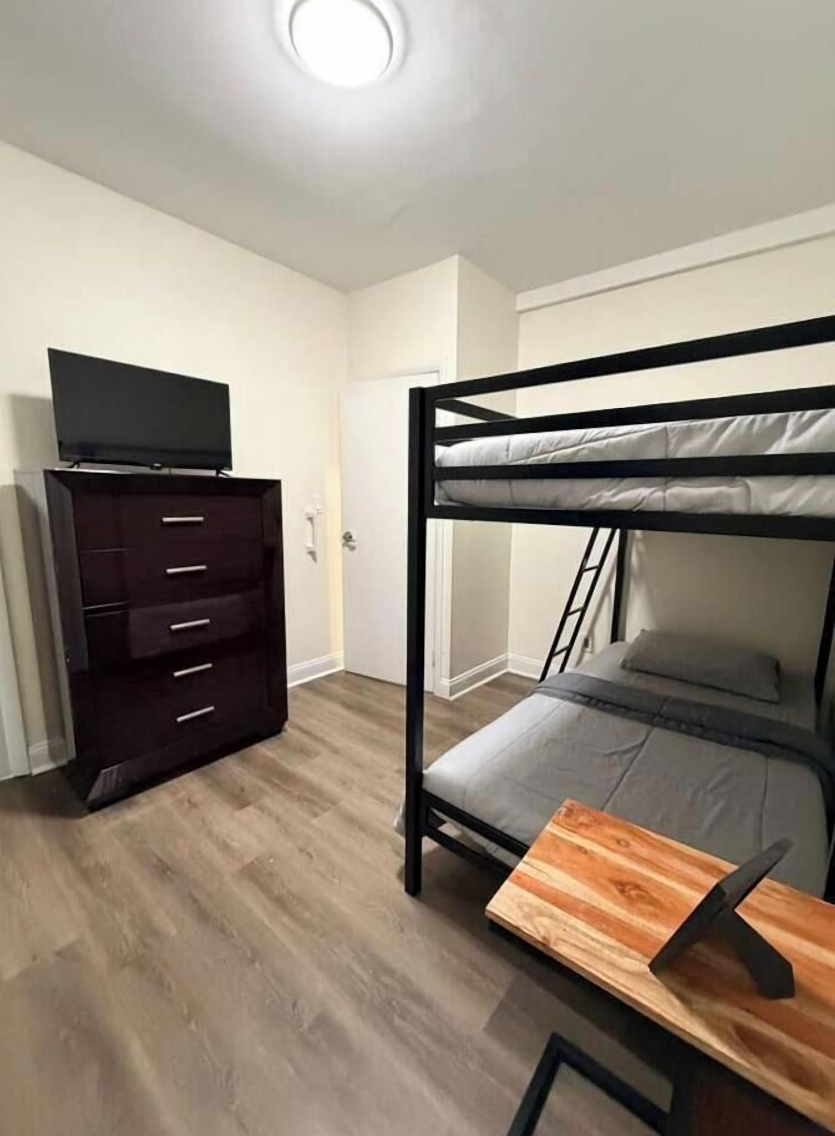 Un lit superposé avec un téléviseur posé sur une commode, une table d’appoint en bois et une porte visible au fond.