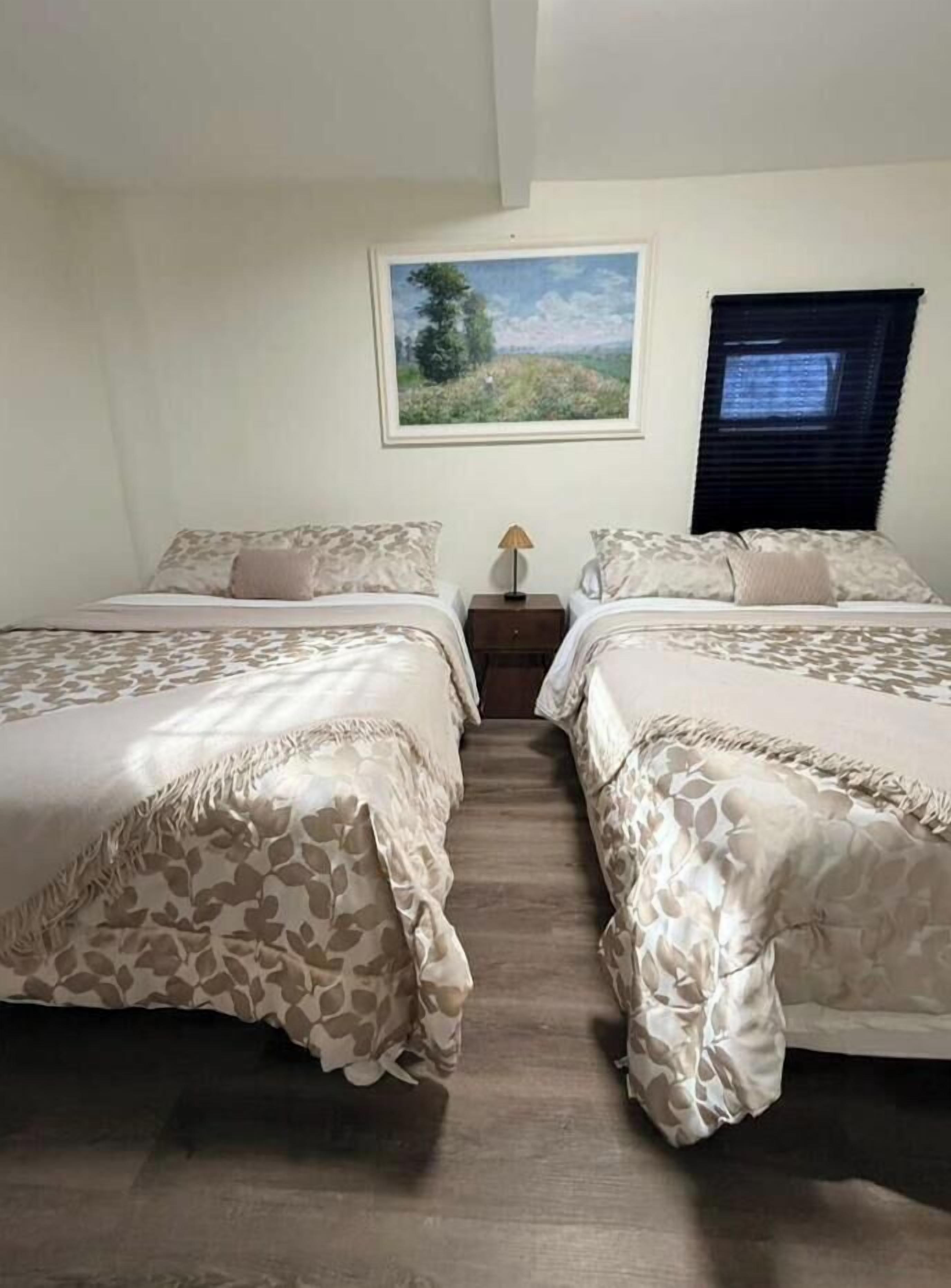 Une chambre avec deux lits, une table de chevet, une lampe, une fenêtre avec des stores et un tableau représentant un paysage accroché au mur.