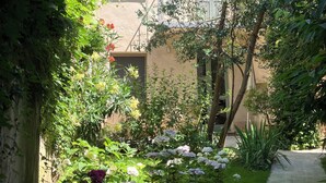 Un patio con un edificio que tiene un balcón, un jardín con diversas plantas y flores, y una pérgola de madera.