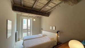 Un dormitorio con dos camas, techo de madera, radiador, escritorio con lámpara y ventana con vista a un balcón.