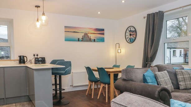 A modern living room with a sofa, a dining table with chairs, a kitchen area with a kettle and toaster, and a large window with curtains. - 6 Hallin Fell (Penrith)