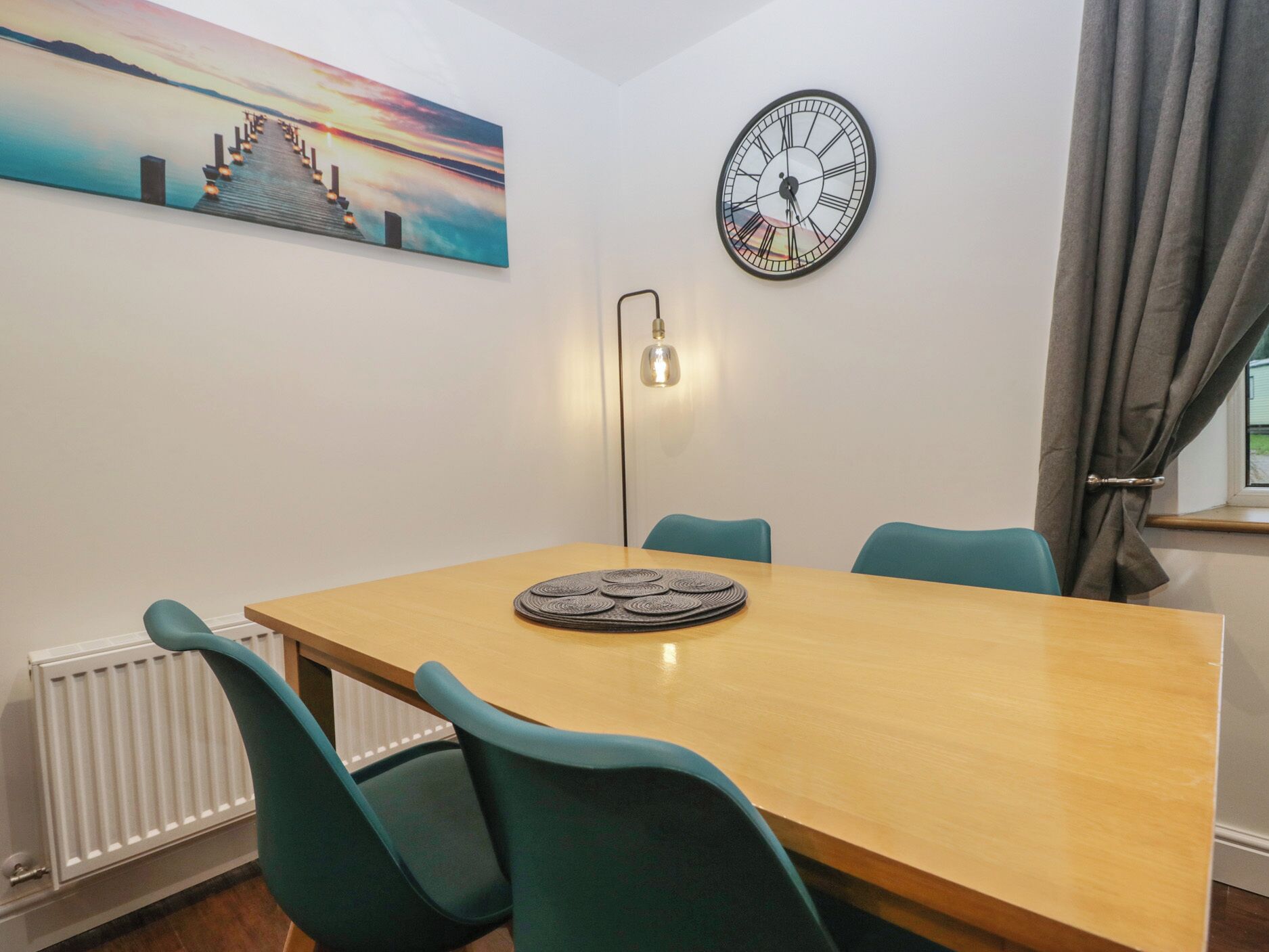A dining area with a wooden table, teal chairs, a wall clock, a painting, and a floor lamp.