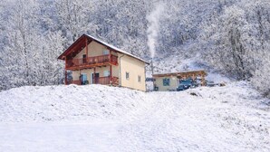Paisaje nevado con una casa de madera que tiene balcón y chimenea humeante.