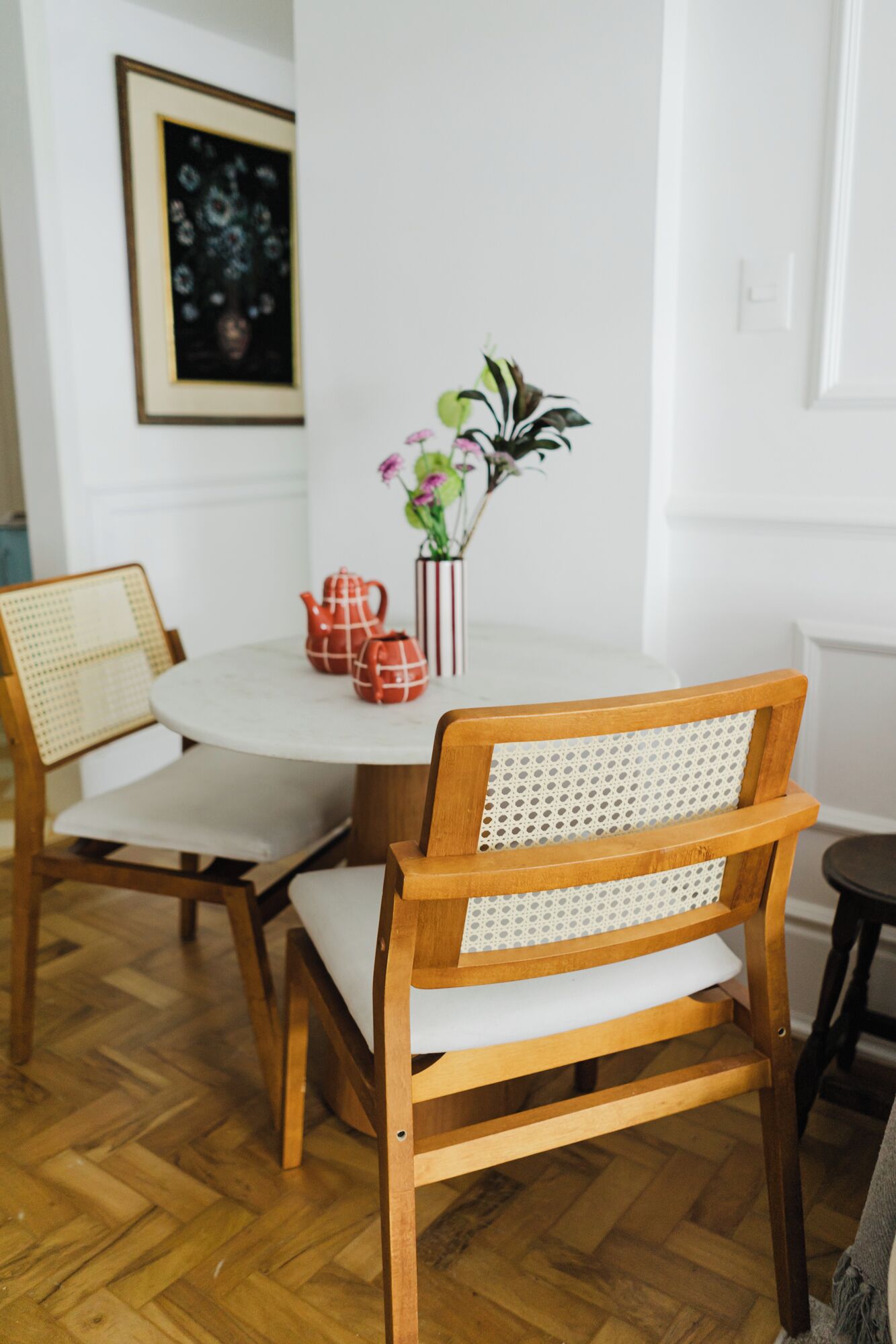 A dining area with a round table, two wooden chairs with woven backs, a vase with flowers, and a red teapot.