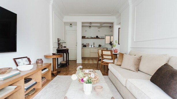 Ein Wohnzimmer mit einer Couch, einem Couchtisch und einem Bücherregal. Im Hintergrund ist eine Küche mit Schränken und einem Spülbecken zu sehen.